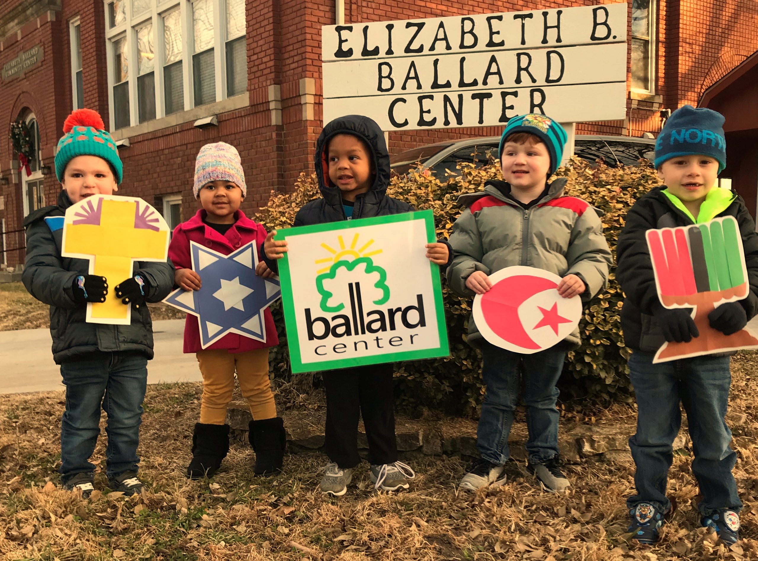 five young children dressed for the cold standing in front of a sign reading Elizabeth B. Ballard Center. Each is holding a religious symbol