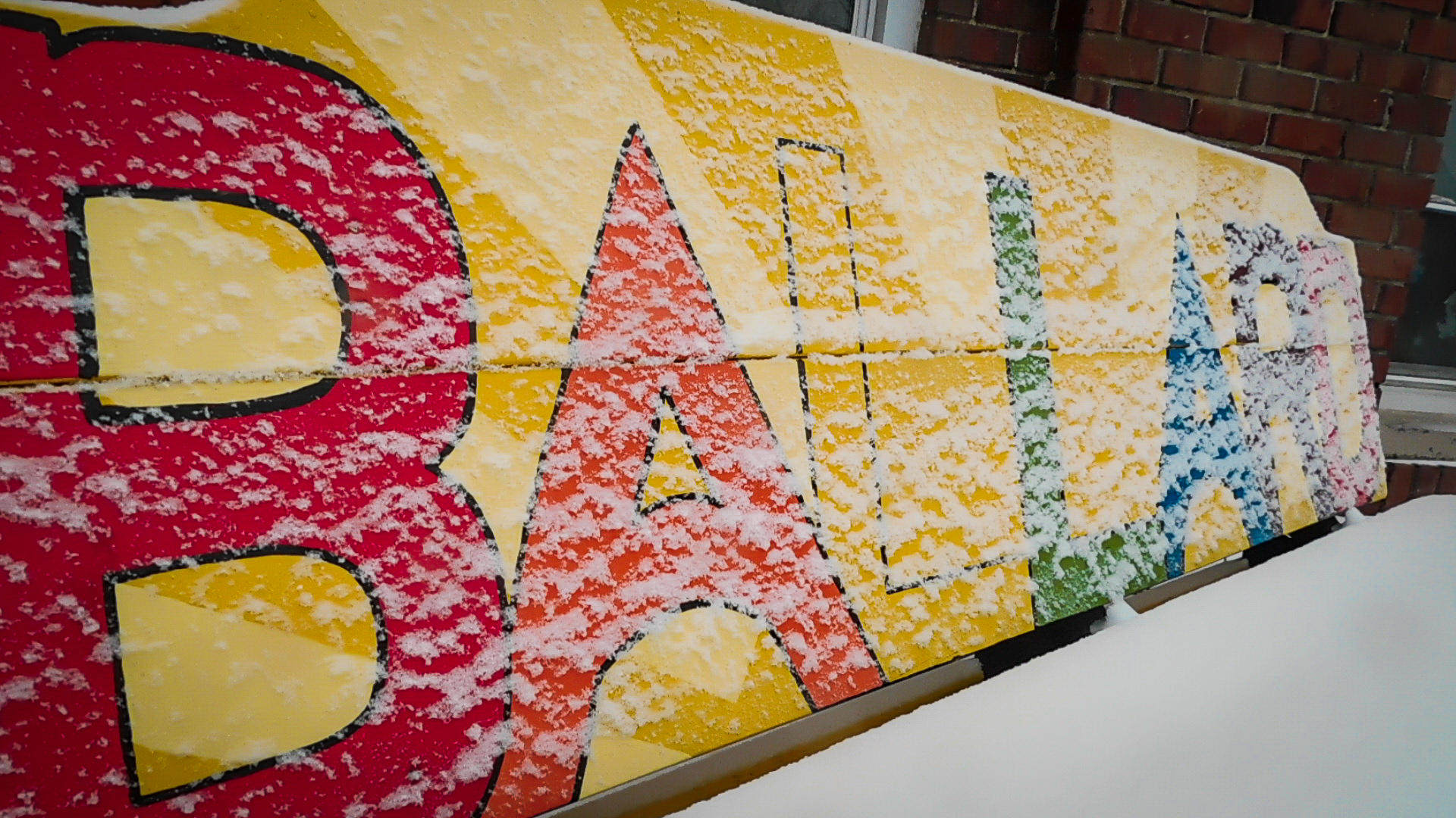 park bench with the word Ballard painted on the back in multi colored letters. it is covered with winter snow