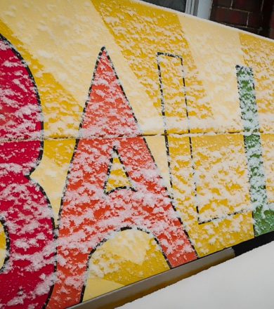 park bench with the word Ballard painted on the back in multi colored letters. it is covered with winter snow