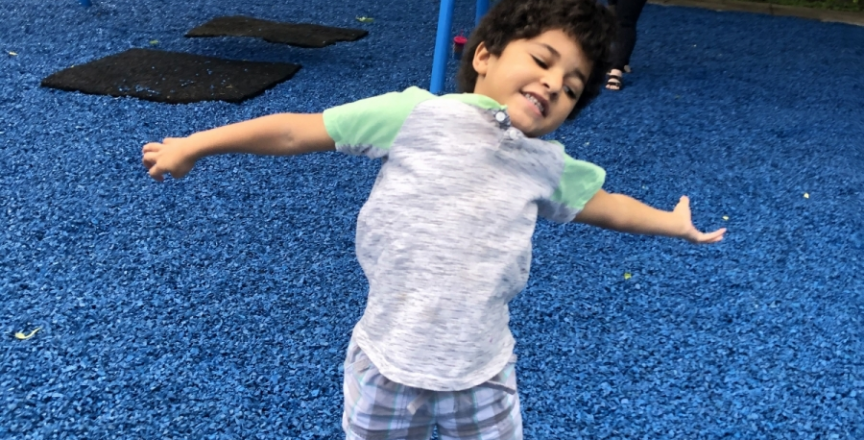 young boy at a playground jumping in the air with his arms out. there is blue playground mulch around him