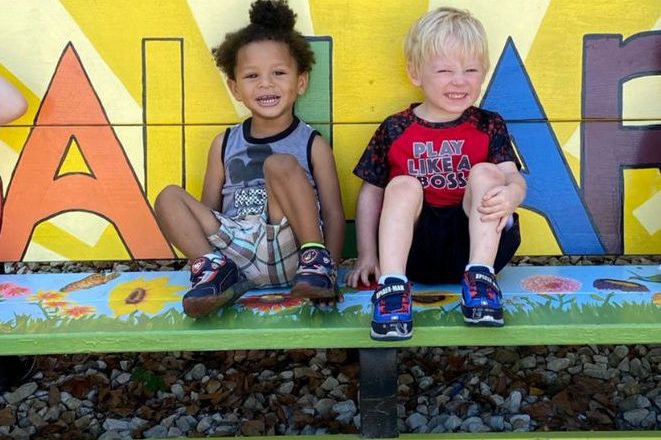 two young children sitting on a multi colored bench. both have their legs up and are smiling