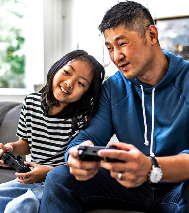 father and daughter sitting on couch holding game consoles