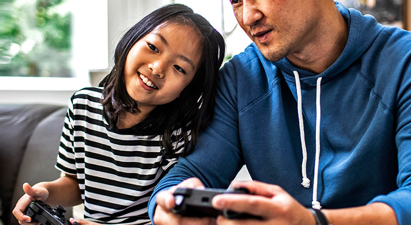 father and daughter sitting on couch holding game consoles