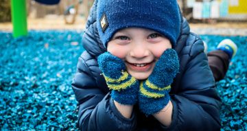 young boy wearing blue coat, gloves and a stocking cap laying down and smiling