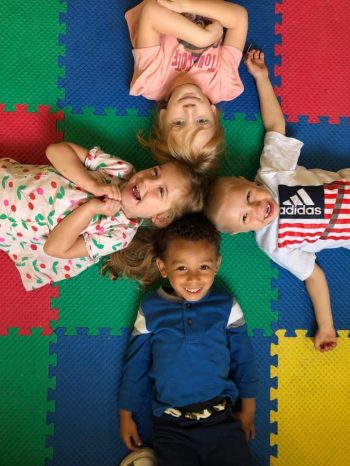 4 young children laying on a multi colored floor smiling at the camera