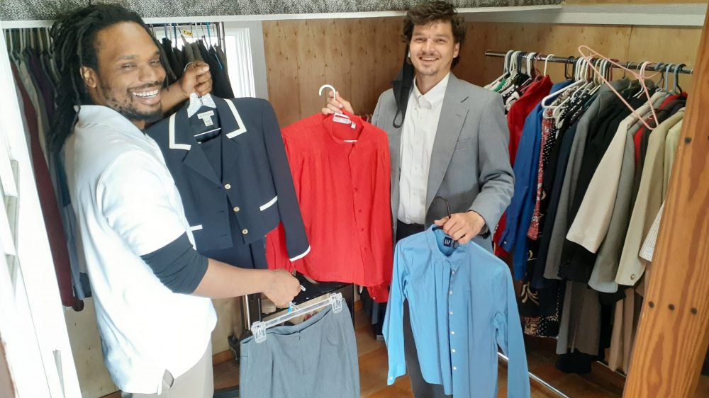 two men holding up suit jackets and shirts in front of racks of shoes and clothes