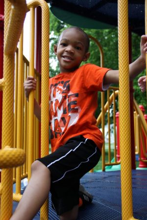 young boy in a orange shirt and black shorts smiling at the camera on a playground set